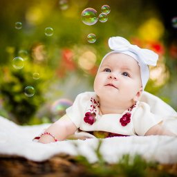Fotografia Damian Wachowiec - Urocza niemowlęca dziewczynka w białej sukience z kokardą na głowie leży na miękkim kocyku, otoczona unoszącymi się bańkami mydlanymi na tle rozmytej zieleni i kwiatów.