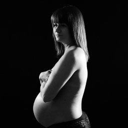 Fotografia Damian Wachowiec - Czarno-białe zdjęcie profilowe kobiety w ciąży, z rękami skrzyżowanymi na piersiach, na czarnym tle.