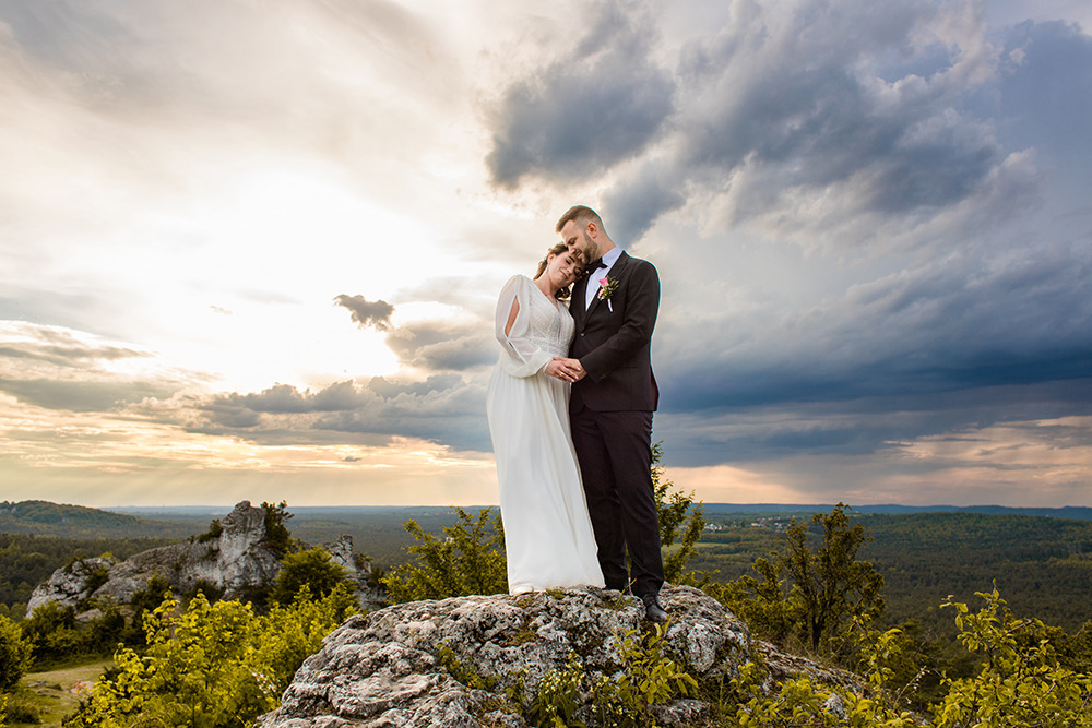 Para młoda stoi na skalistym wzgórzu z widokiem na zalesioną dolinę pod dramatycznym, zachodzącym słońcem. Panna młoda w zwiewnej sukni z długimi rękawami opiera głowę o ramię pana młodego...
