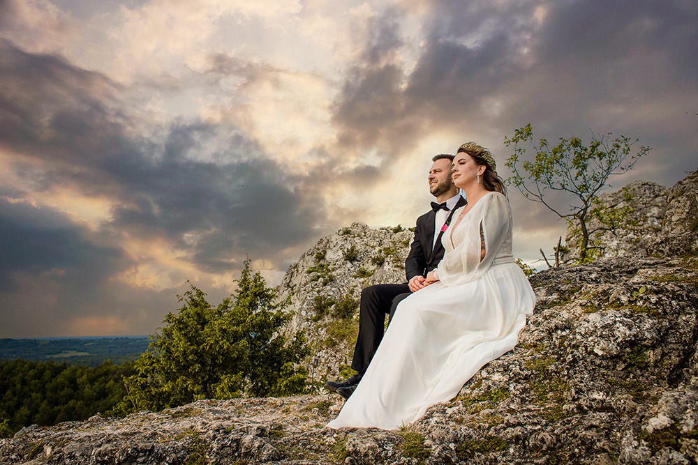 Para młoda siedzi na skalistym wzgórzu podczas sesji plenerowej, w tle dramatyczne chmury oświetlone zachodzącym słońcem.