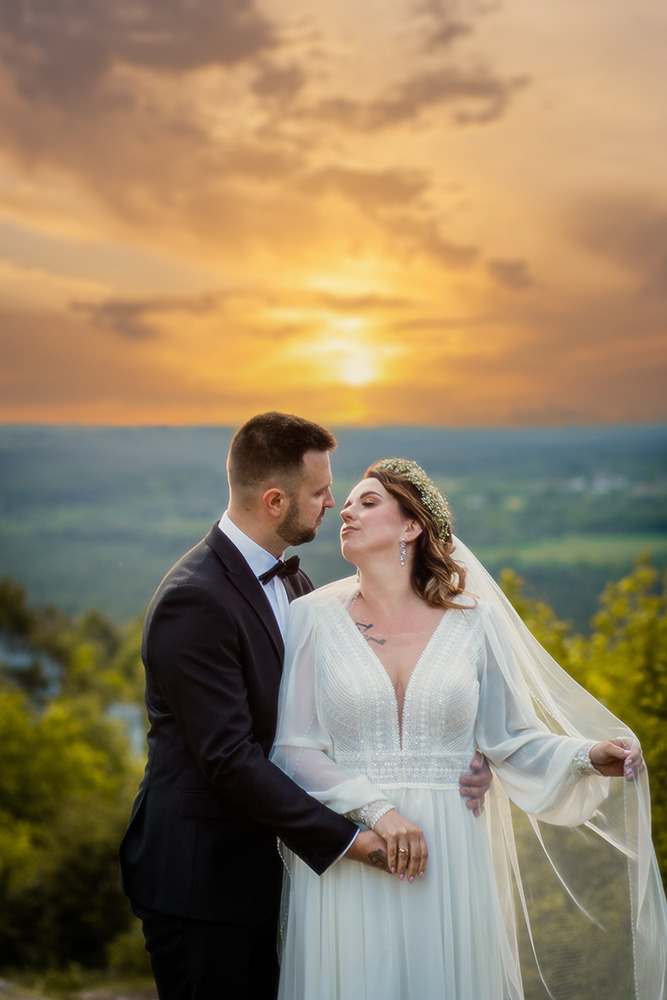Para młoda w eleganckich strojach, delikatny uśmiech, na tle zachodzącego słońca i malowniczego krajobrazu zalesionych wzgórz.