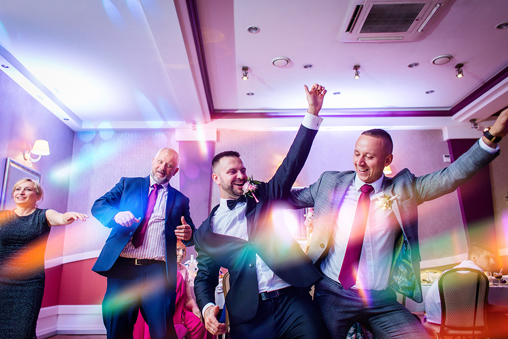 Rozbawieni goście weselni tańczą na parkiecie oświetlonym kolorowymi światłami, w tle widoczni siedzący goście przy stolikach.