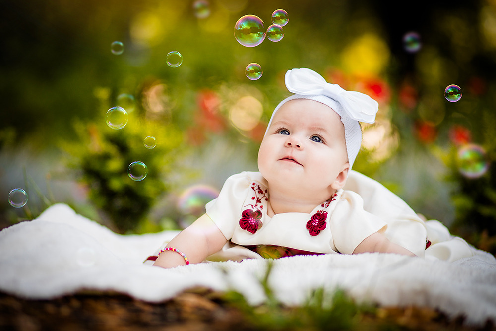 Urocza niemowlęca dziewczynka w białej sukience z kokardą na głowie leży na miękkim kocyku, otoczona unoszącymi się bańkami mydlanymi na tle rozmytej zieleni i kwiatów.