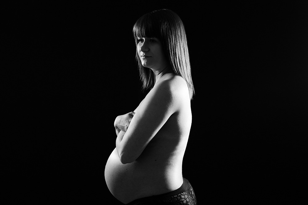Czarno-białe zdjęcie profilowe kobiety w ciąży, z rękami skrzyżowanymi na piersiach, na czarnym tle.