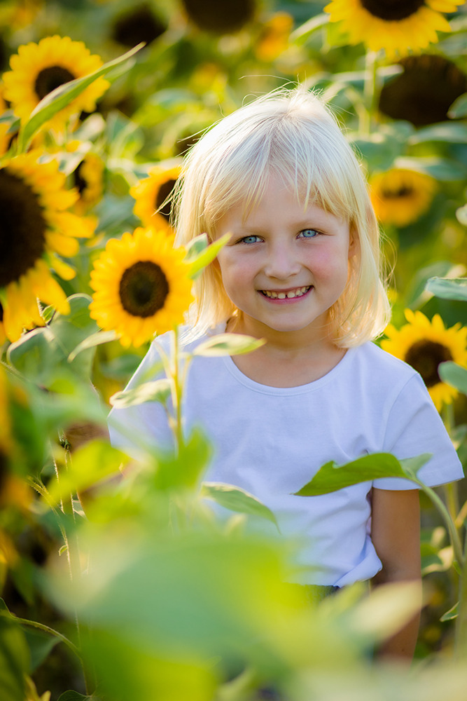 Portret uśmiechniętej, jasnowłosej dziewczynki z niebieskimi oczami w polu słoneczników w Łodzi, ubrana w białą koszulkę, z delikatnym, naturalnym makijażem.