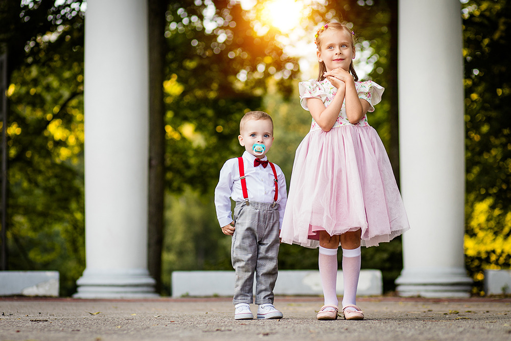 Urocze rodzeństwo: chłopiec z czerwoną muszką i szelkami oraz dziewczynka w różowej sukience, pozują na tle klasycznych kolumn i zieleni.