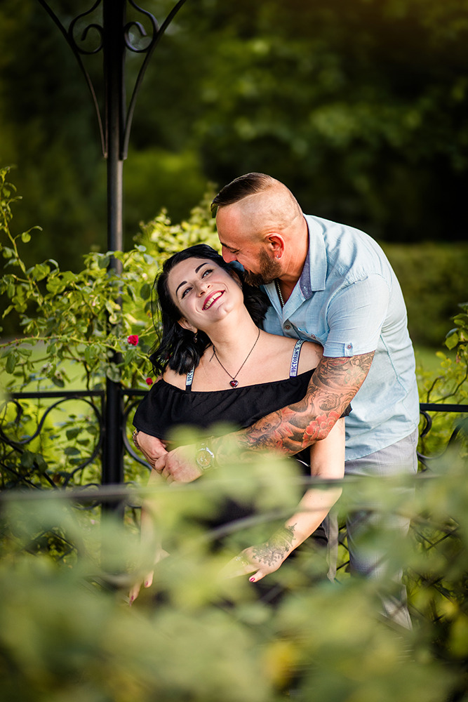 Uśmiechnięta kobieta w czarnej sukience, obejmowana przez mężczyznę z tatuażami na tle zieleni w parku.