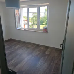 Pusty pokój z brązowymi panelami podłogowymi, świeżo pomalowanymi białymi ścianami, oknem z widokiem na zieleń i puszką farby na parapecie, widok zza otwartych drzwi.