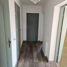 Korytarz po remoncie z trzema parami drzwi w odcieniach zieleni, podłoga z paneli imitujących drewno, białe ściany i geometryczna lampa sufitowa.