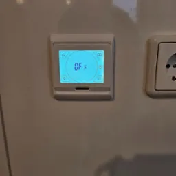 Biały termostat pokojowy zamontowany na kafelkach ściennych, wyświetlający napis 'OFF' na niebieskim ekranie LCD, obok gniazdka elektrycznego