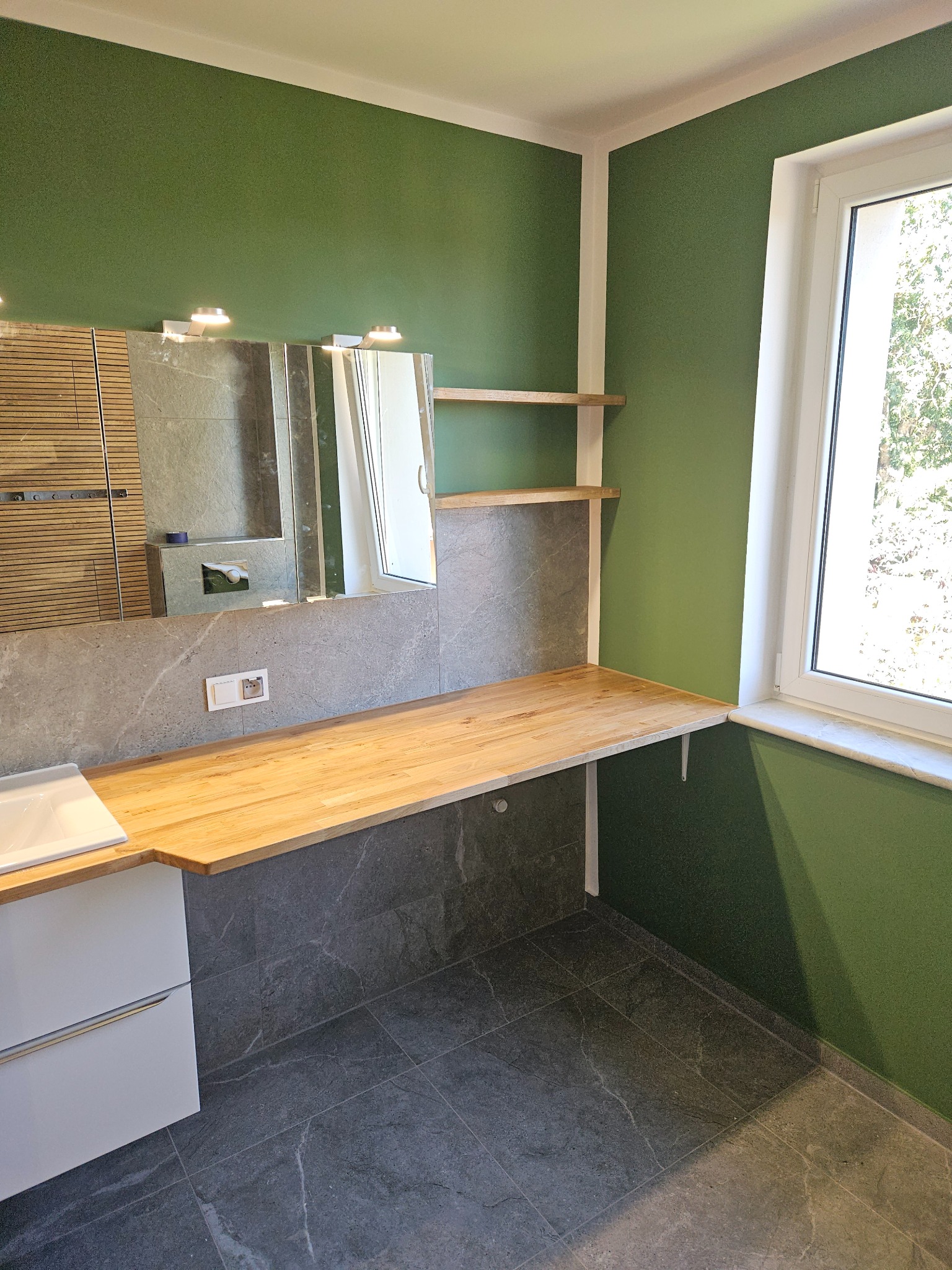 Nowoczesna łazienka z zielonymi ścianami, szarą płytką imitującą kamień, drewnianym blatem i białymi szafkami pod umywalką, oświetlona światłem dziennym z okna.
