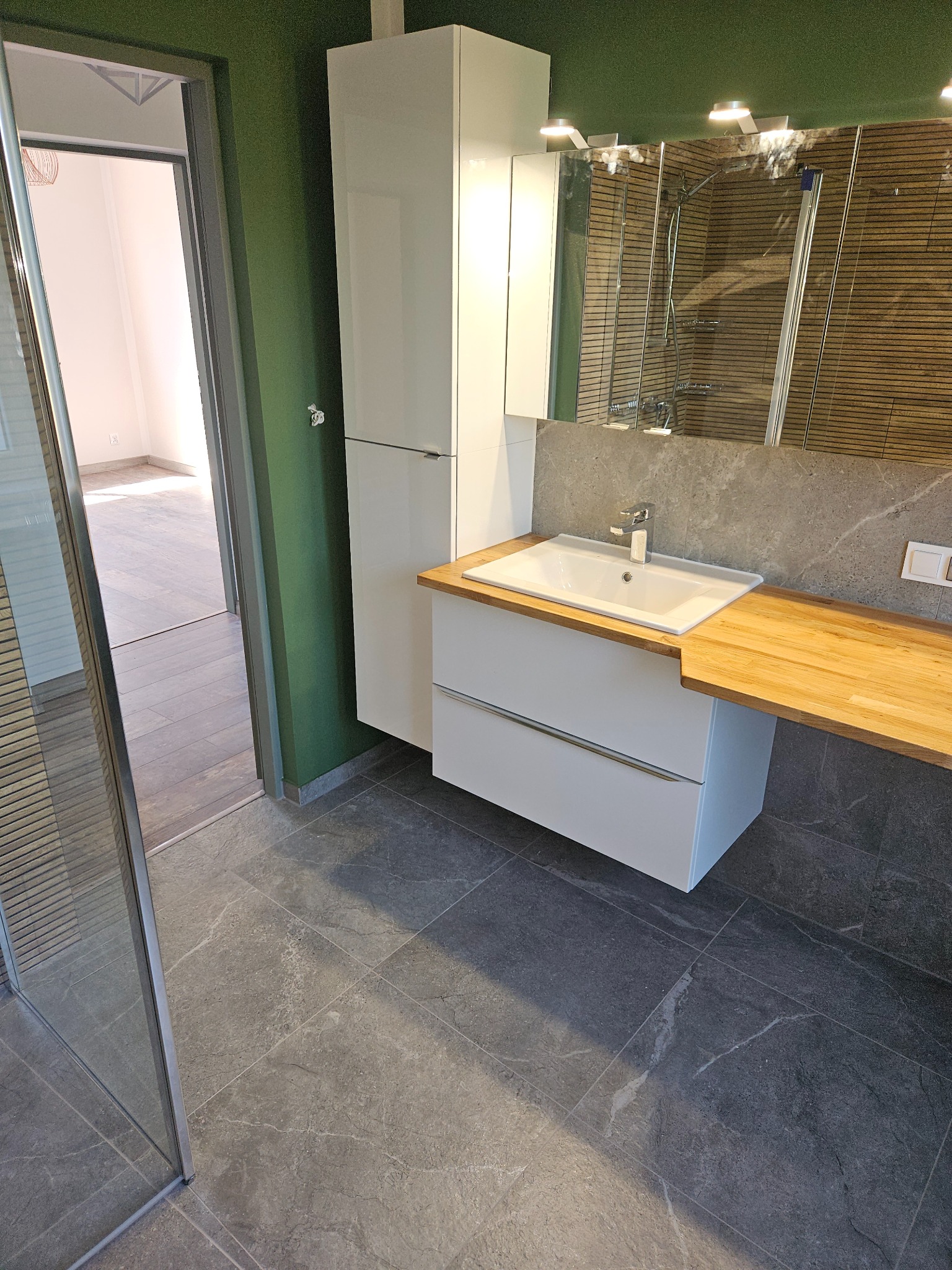 Nowoczesna łazienka z szarą płytką podłogową imitującą kamień, białą szafką pod umywalką z drewnianym blatem, wysoką białą szafką wiszącą i kabiną prysznicową z widocznymi poziomymi panelami.