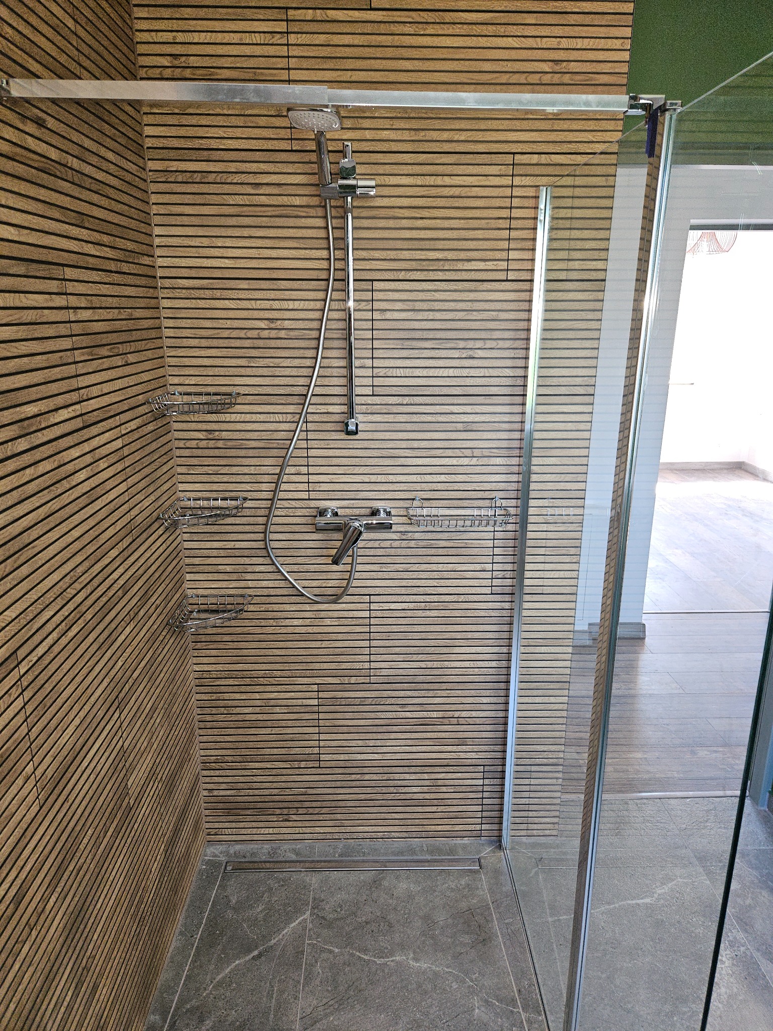 Kabina prysznicowa z drewnianą okładziną ścienną, szklanymi drzwiami i odpływem liniowym w podłodze z szarego kamienia.