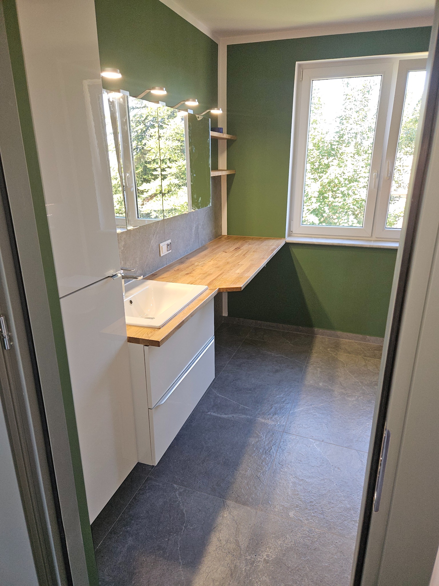 Nowoczesna łazienka w Czarnej Dąbrówce z zielonymi ścianami, szarą podłogą z płytek imitujących kamień, drewnianym blatem i białą szafką z umywalką, widok zza uchylonych drzwi.