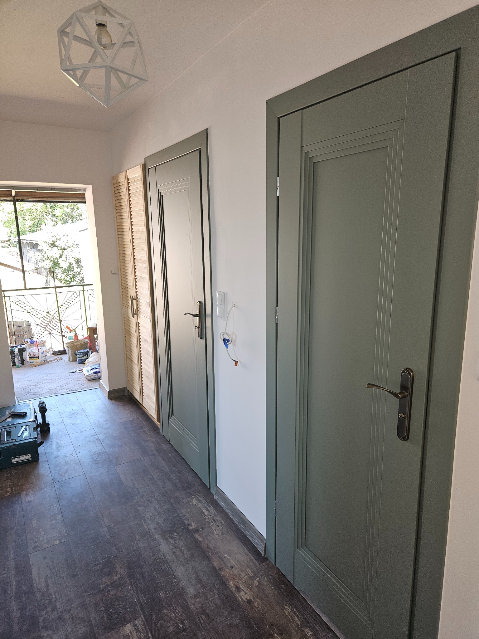 Wnętrze korytarza z trzema nowymi, oliwkowymi drzwiami z brązowymi klamkami, ciemna podłoga imitująca drewno, widoczne narzędzia i materiały budowlane na balkonie.