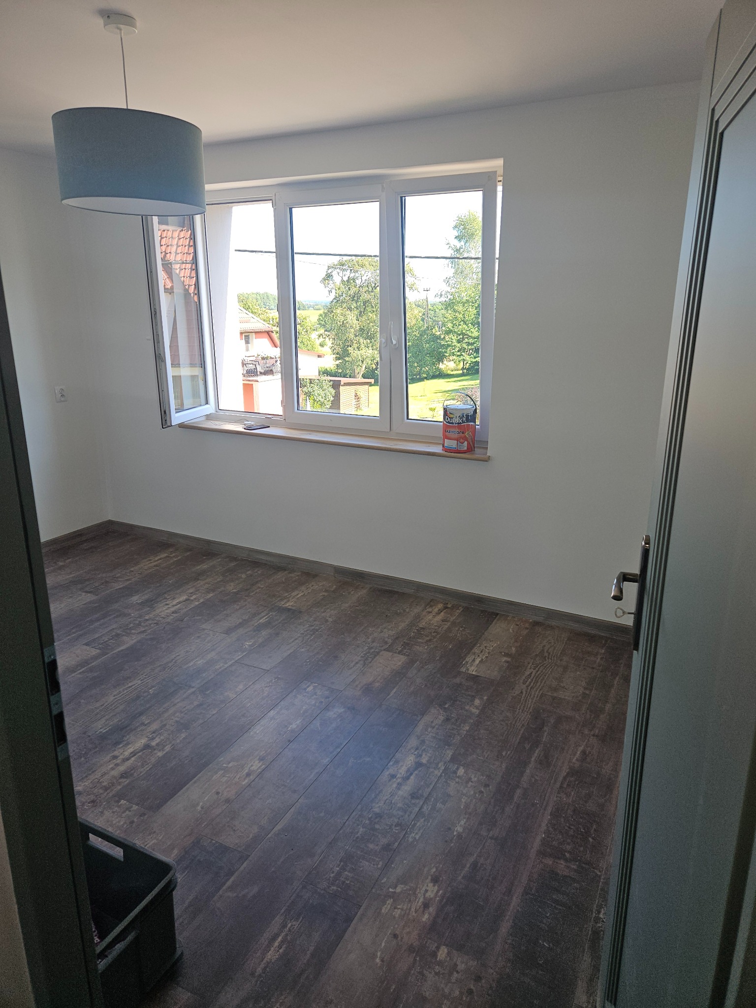 Pusty pokój z brązowymi panelami podłogowymi, świeżo pomalowanymi białymi ścianami, oknem z widokiem na zieleń i puszką farby na parapecie, widok zza otwartych drzwi.