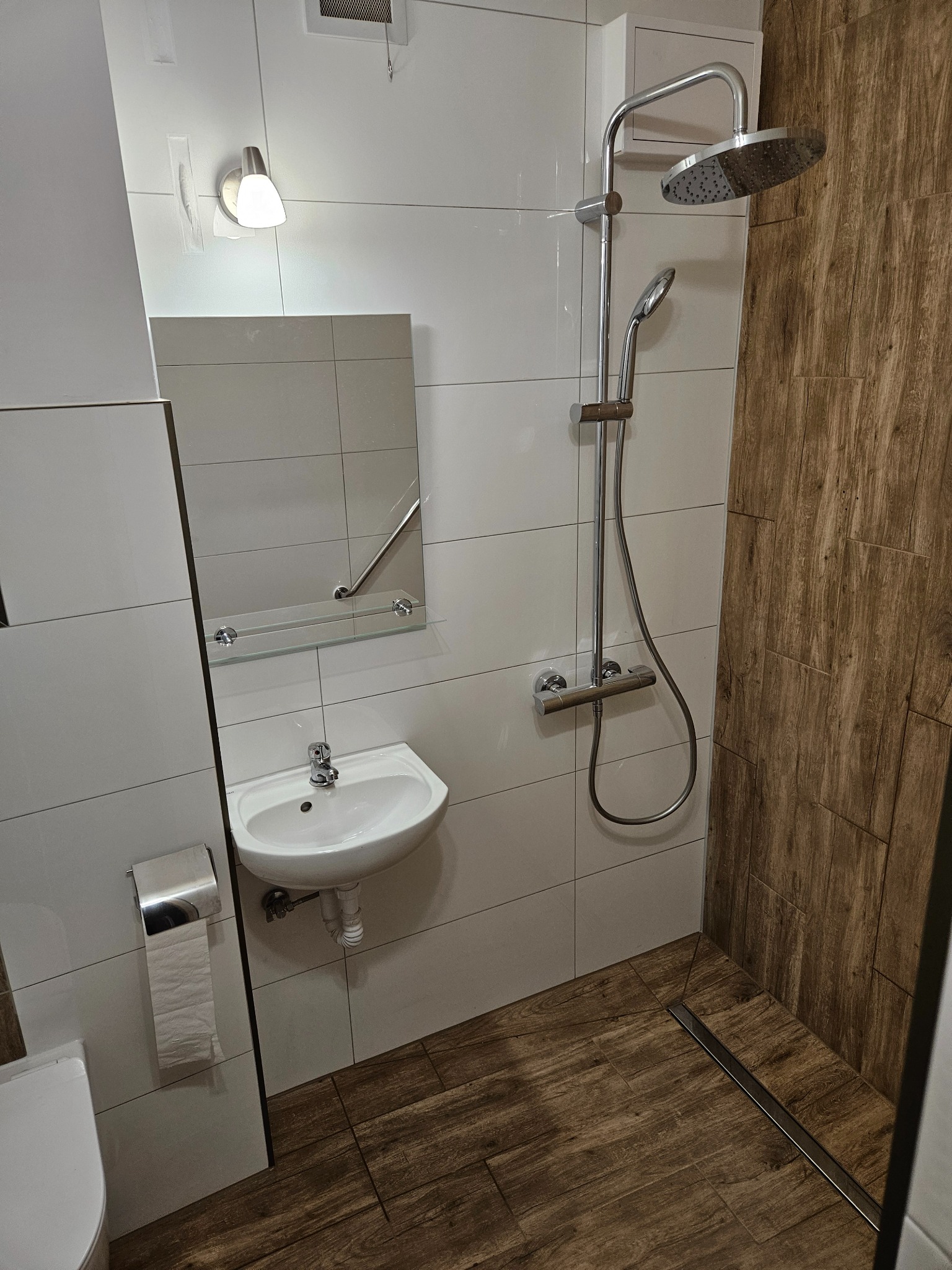 Nowoczesna łazienka z kabiną prysznicową typu walk-in, wykończona białymi płytkami i panelami imitującymi drewno, z widocznym odpływem liniowym i umywalką z lustrem.