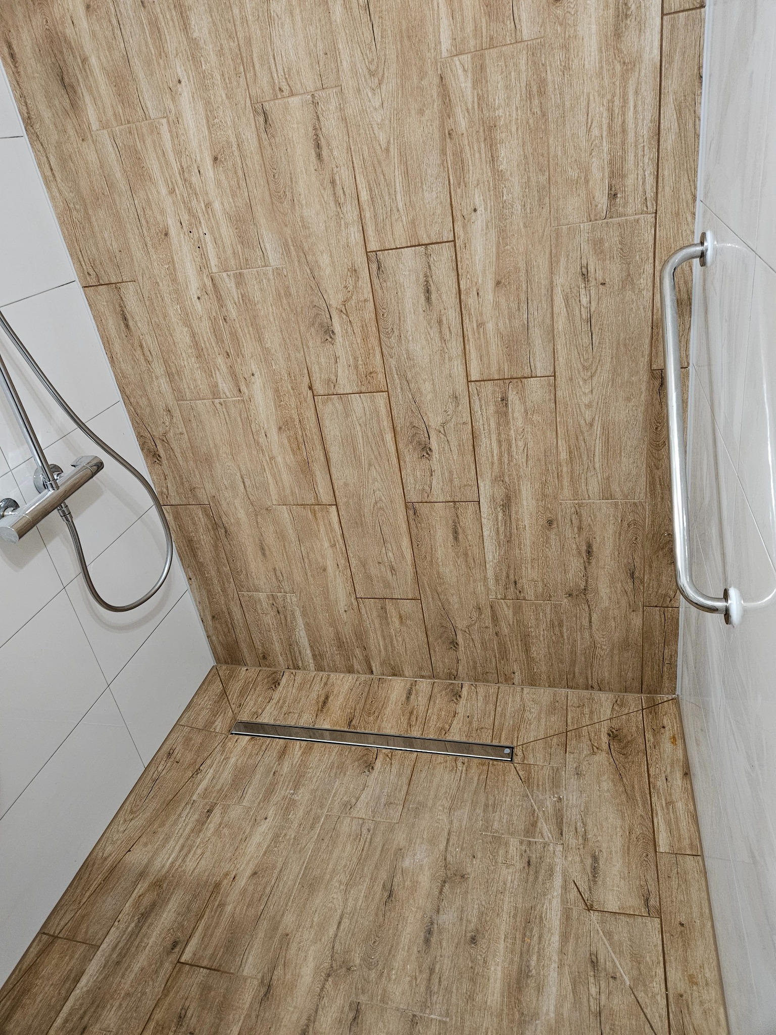 Kabina prysznicowa wykończona płytkami imitującymi drewno, z odpływem liniowym i chromowaną armaturą.