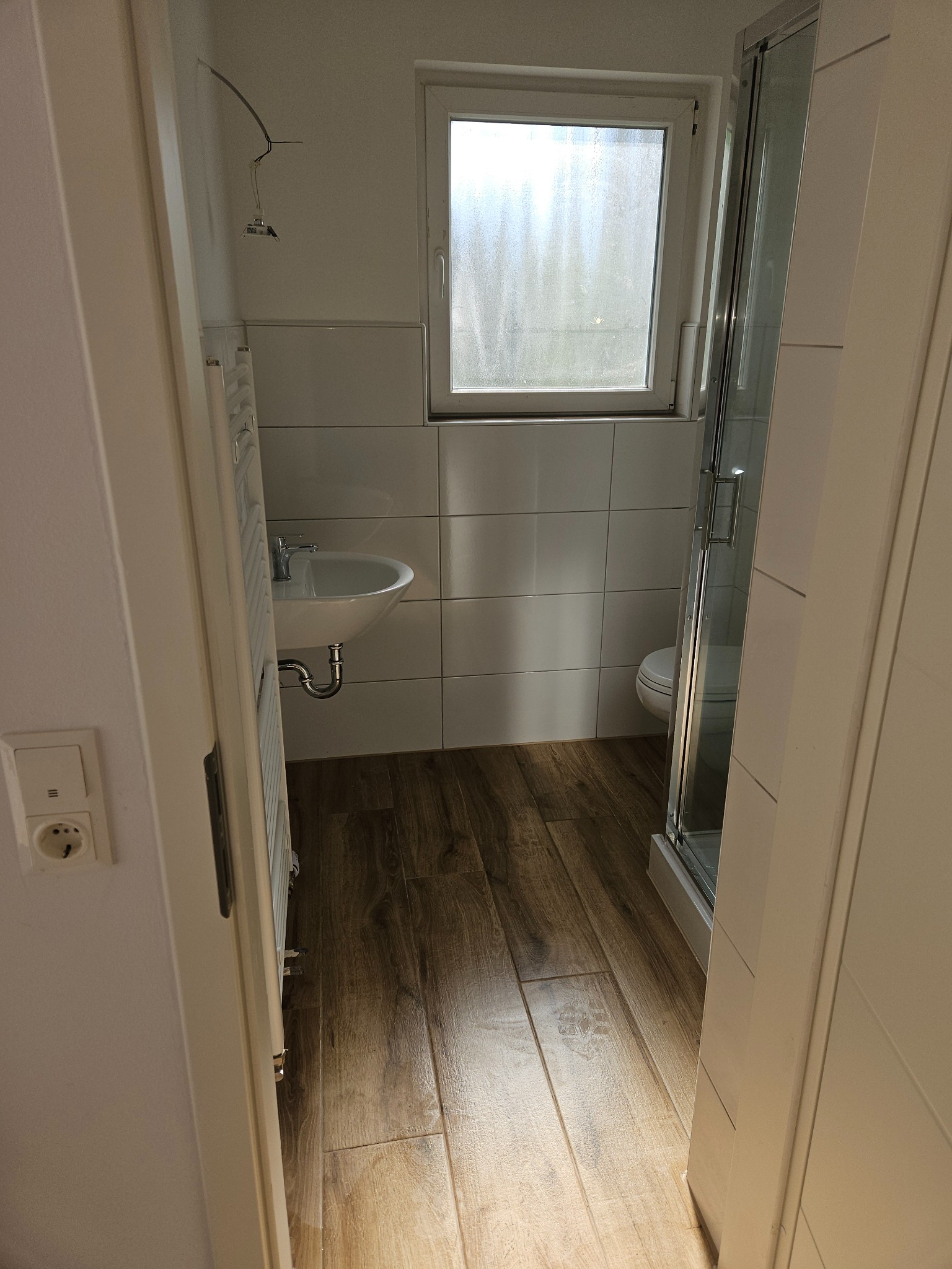 Widok na łazienkę z brązowymi panelami podłogowymi, białą umywalką i kabiną prysznicową, widoczny przez otwarte drzwi, z niedokończonym podłączeniem oświetlenia sufitowego.