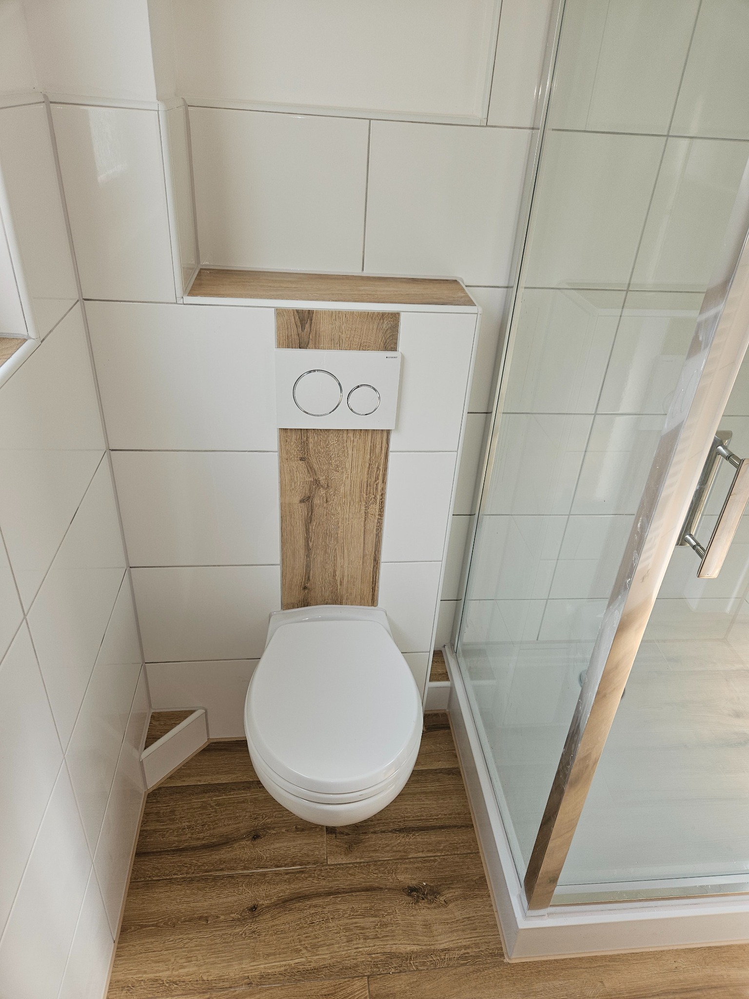 Nowoczesna łazienka z białymi płytkami, drewnopodobną podłogą i zabudową WC z drewnianym akcentem oraz kabiną prysznicową ze srebrnym uchwytem, widok z góry.