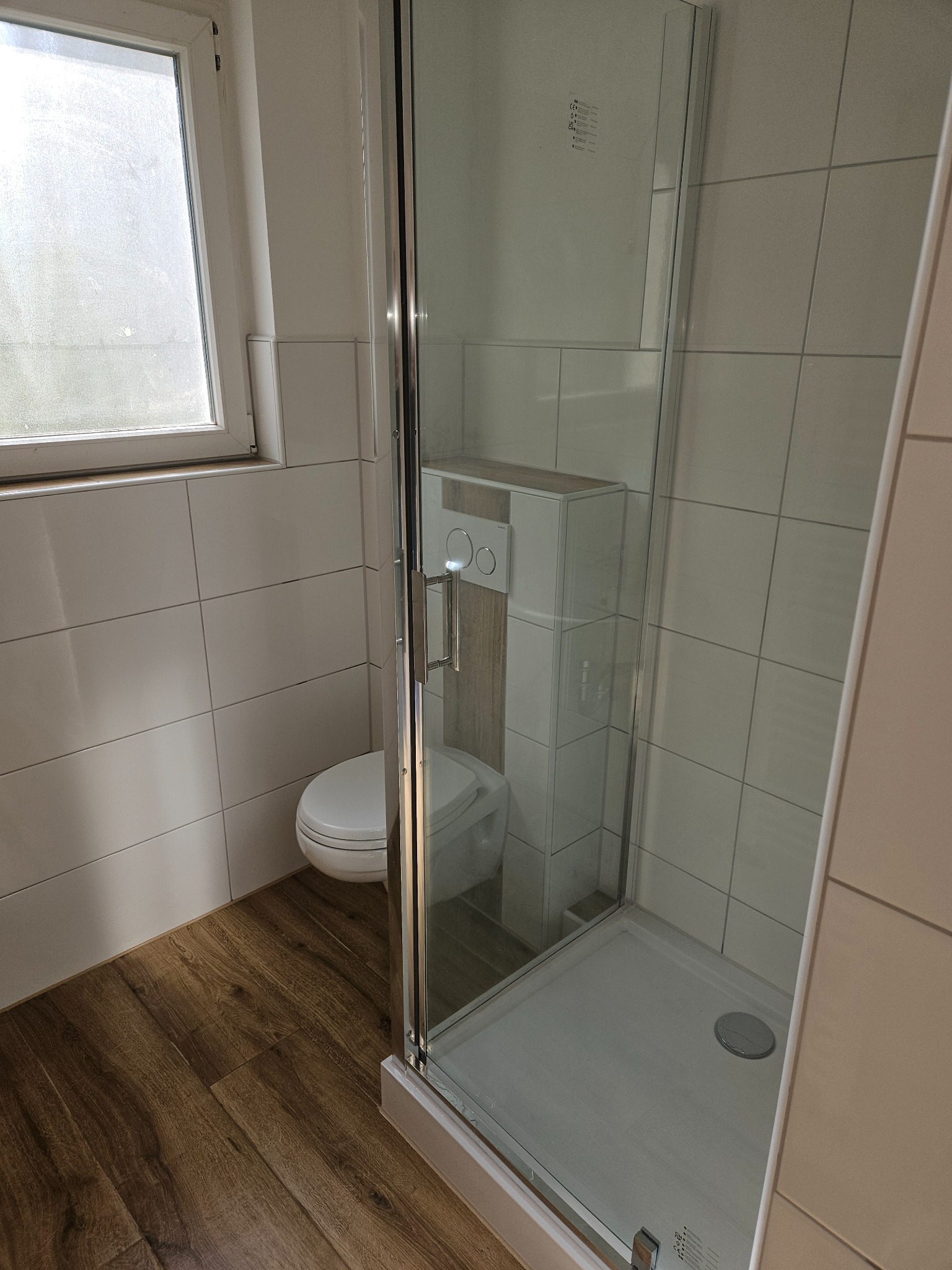 Nowoczesna kabina prysznicowa ze szklanymi drzwiami, widoczny fragment podwieszanej toalety z drewnianym akcentem, jasne płytki na ścianach i drewnopodobna podłoga.