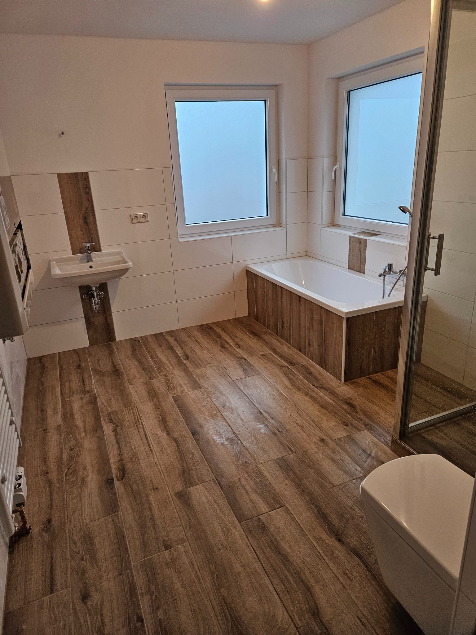 Nowoczesna łazienka z drewnopodobną podłogą, białą wanną obudowaną panelami w kolorze drewna, umywalką i kabiną prysznicową z transparentnego szkła.