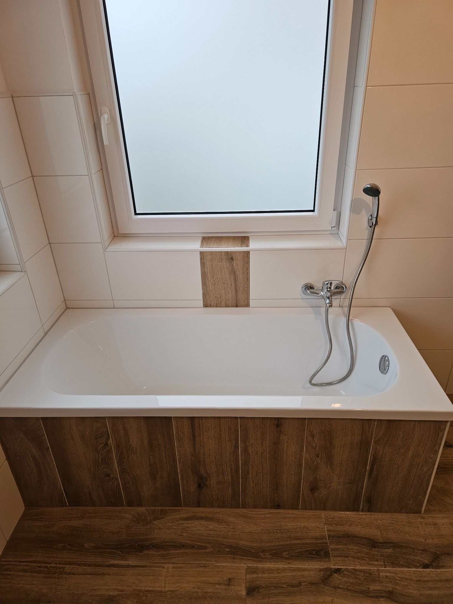 Biała wanna obudowana panelami imitującymi drewno, z baterią i słuchawką prysznicową, pod oknem dachowym w łazience wyłożonej jasnymi płytkami.