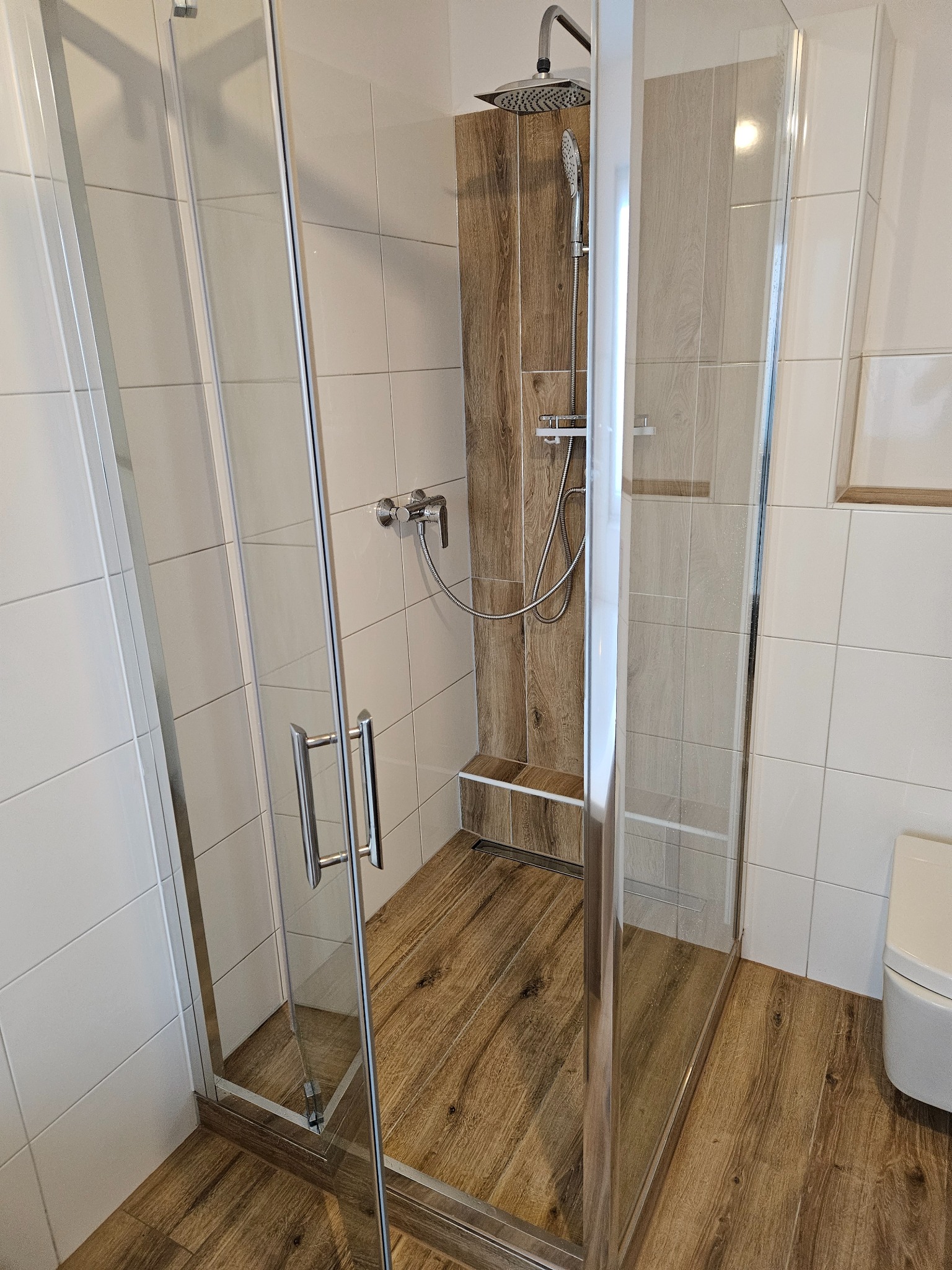 Kabina prysznicowa ze szklanymi drzwiami, wykończona białymi płytkami i panelami imitującymi drewno, z deszczownicą i baterią termostatyczną.