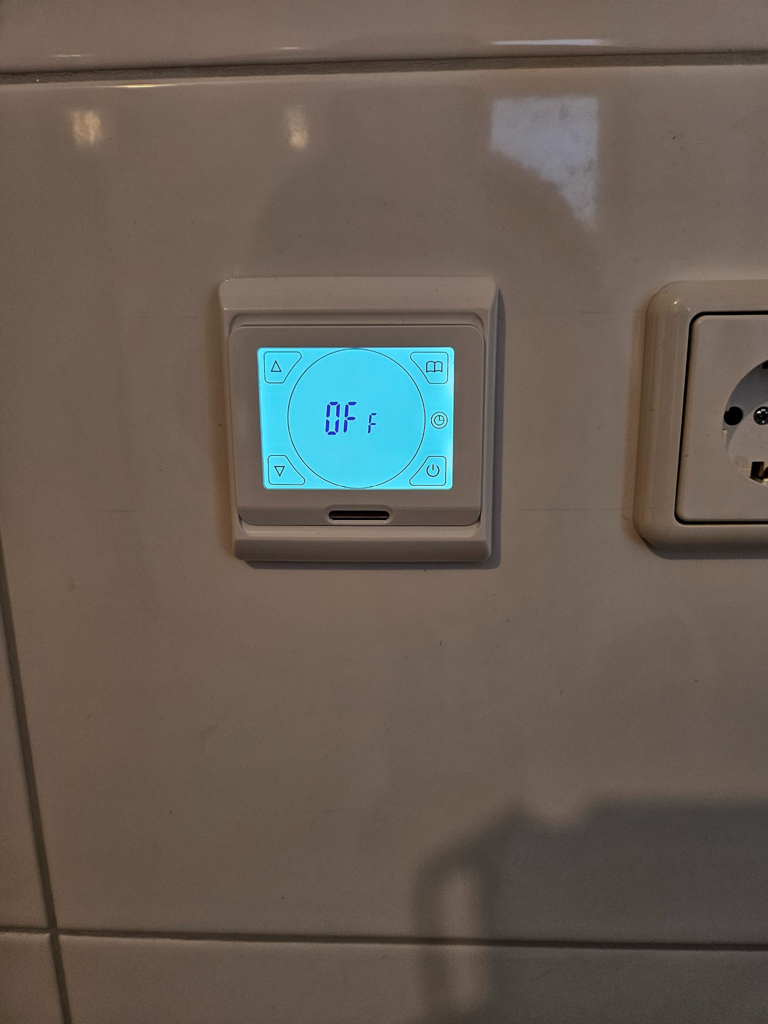 Biały termostat pokojowy zamontowany na kafelkach ściennych, wyświetlający napis 'OFF' na niebieskim ekranie LCD, obok gniazdka elektrycznego