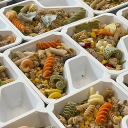 Gramatura Catering - Porcje makaronu z kurczakiem i warzywami w białych, plastikowych pojemnikach z przegródkami, ułożone obok siebie, makaron w różnych kolorach i kształtach.