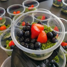 Gramatura Catering - Deser w przezroczystym pojemniku z pokruszoną zieloną posypką, borówkami i kawałkami truskawek, trzymany w rękawicy.