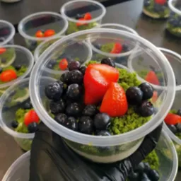 Deser w przezroczystym pojemniku z pokruszoną zieloną posypką, borówkami i kawałkami truskawek, trzymany w rękawicy.