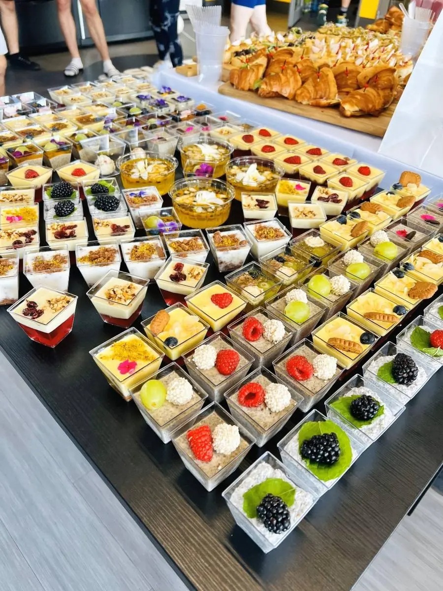 Różnorodne desery w przezroczystych pojemnikach, w tym musy owocowe, puddingi chia i ciastka, ułożone na stole podczas cateringu.