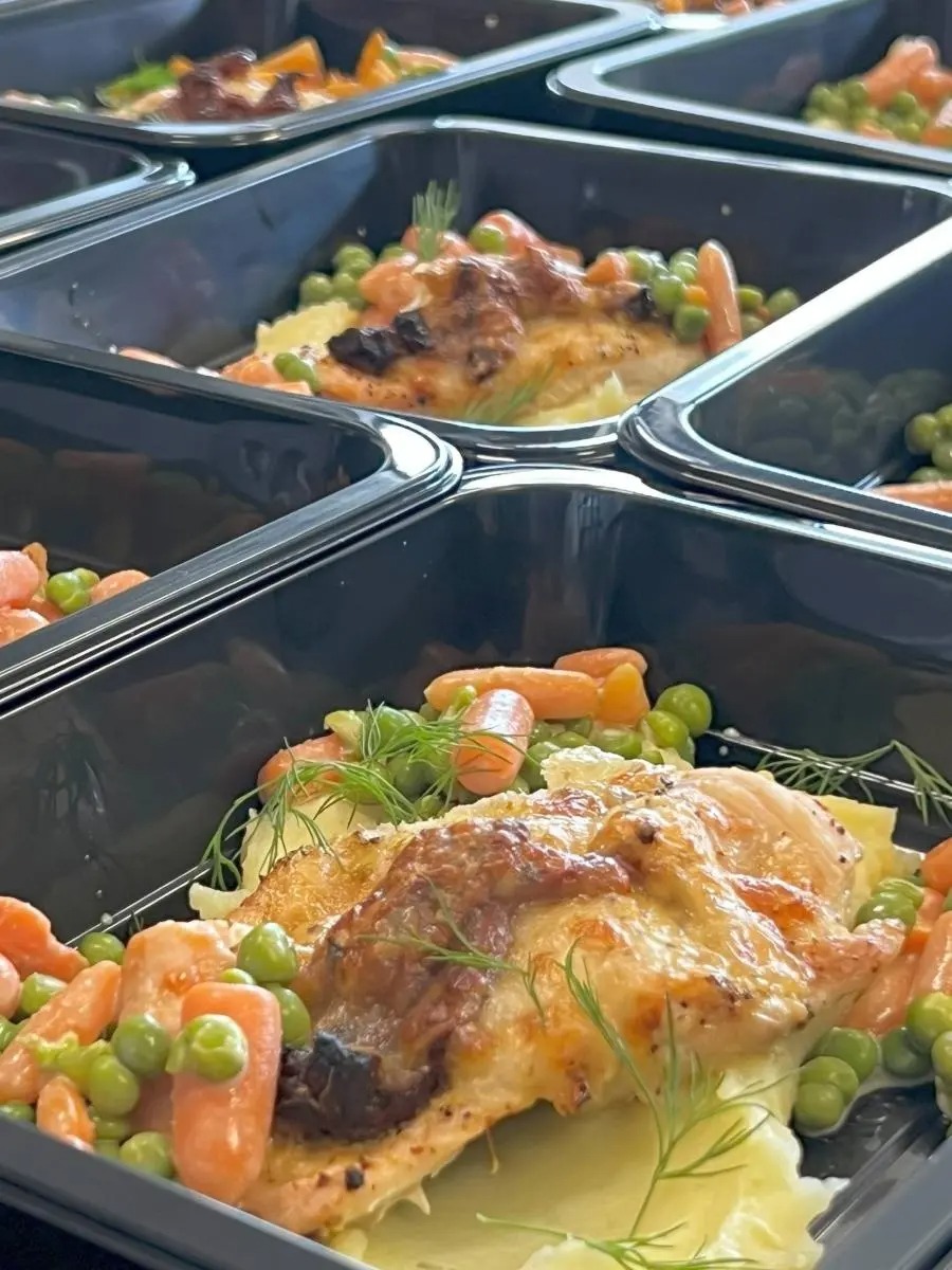 Porcje pieczonego kurczaka z puree ziemniaczanym, marchewką i groszkiem w czarnych pojemnikach cateringowych, udekorowane koperkiem.