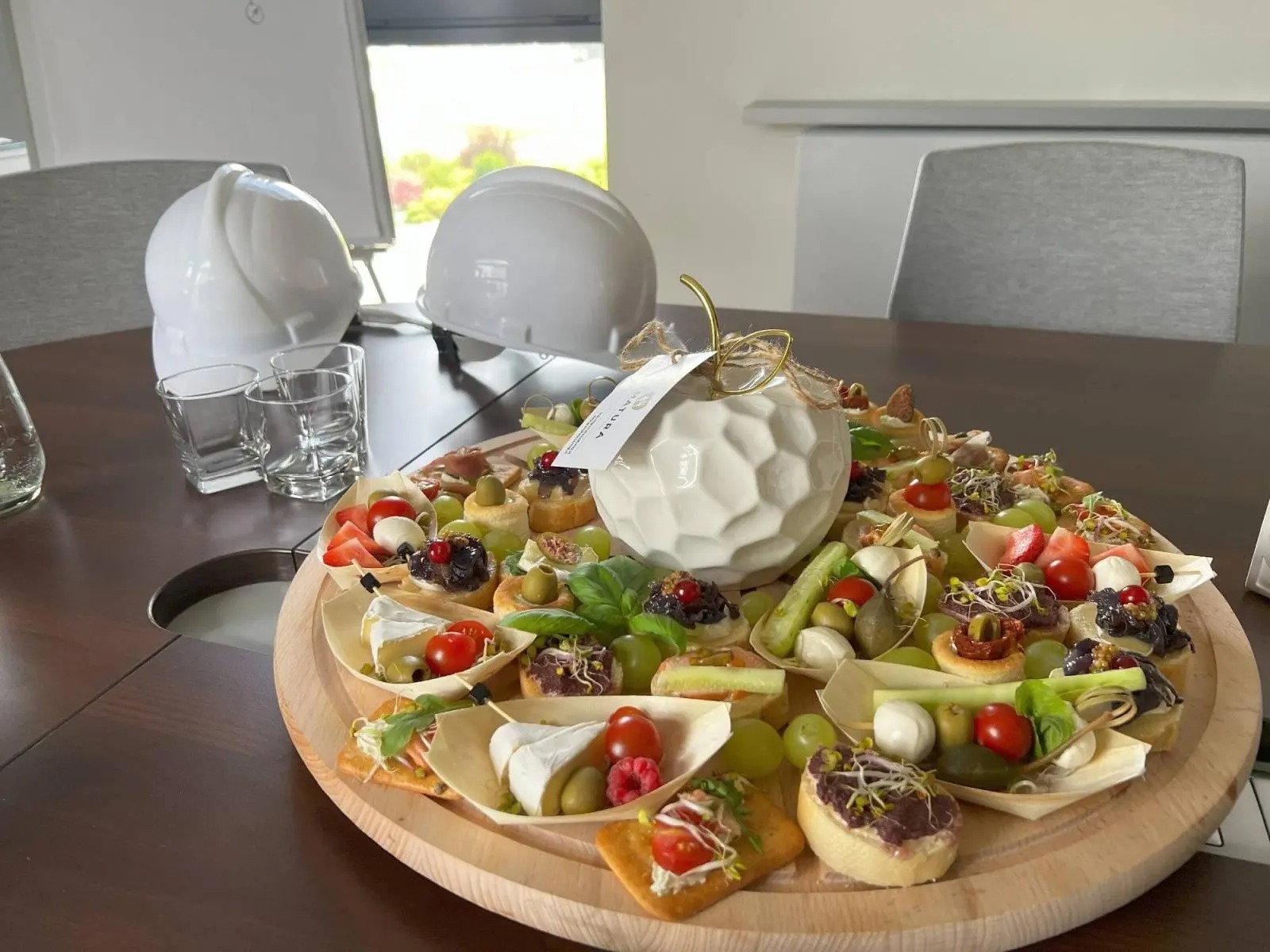 Drewniana taca z różnorodnymi przekąskami, w tle dwa białe kaski budowlane i szklanki, co sugeruje catering dla firmy budowlanej w Raciborzu.