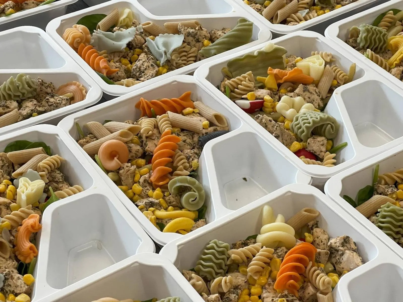 Porcje makaronu z kurczakiem i warzywami w białych, plastikowych pojemnikach z przegródkami, ułożone obok siebie, makaron w różnych kolorach i kształtach.