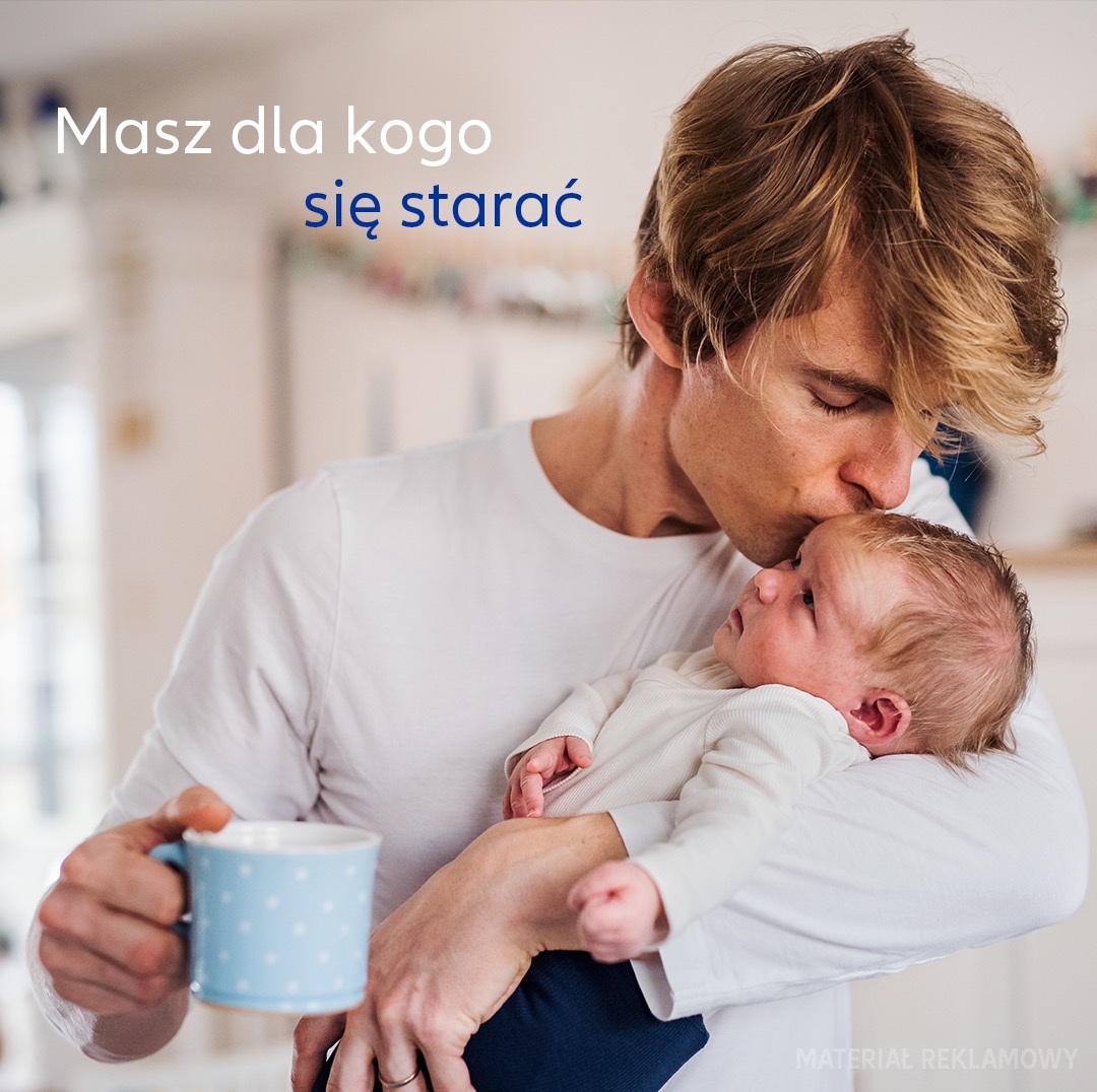 Młody ojciec całuje w czoło noworodka, trzymając w drugiej ręce błękitny kubek w białe kropki, w tle jasne wnętrze mieszkania.