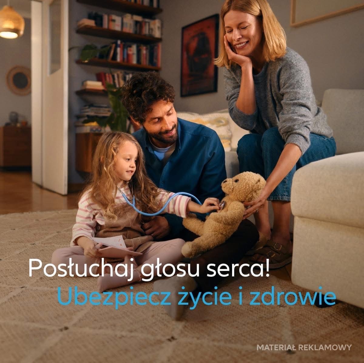 Rodzina w salonie: córka bada stetoskopem pluszowego misia, ojciec obserwuje z uśmiechem, matka siedzi na kanapie z ręką na policzku, w tle regał z książkami.