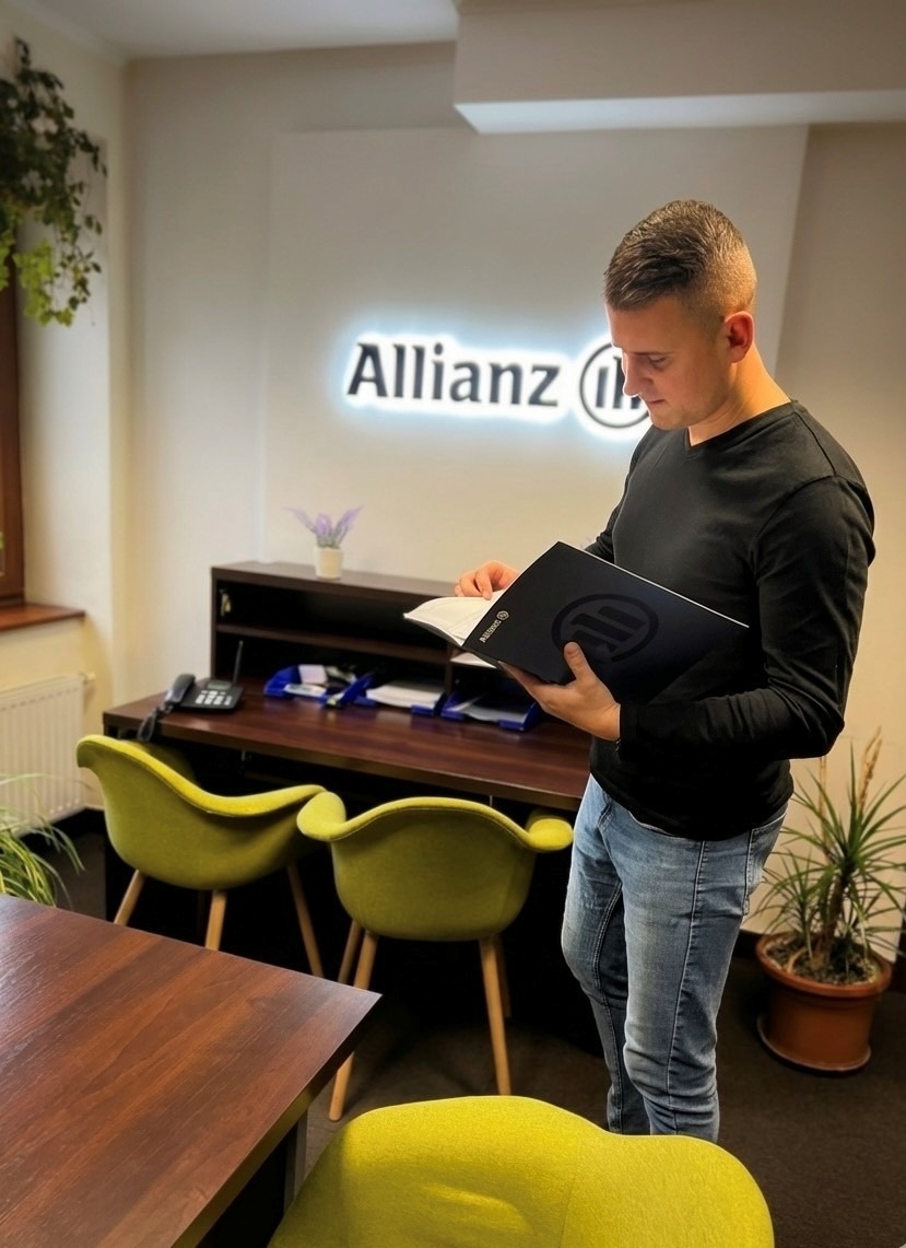 Mężczyzna w biurze Allianz przegląda dokumenty, w tle recepcja z logo firmy, zielone krzesła i rośliny doniczkowe. Stonowana kolorystyka wnętrza.