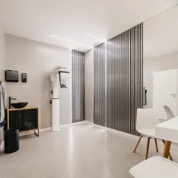 Wnętrze nowoczesnej kliniki stomatologicznej z aparaturą RTG, umywalką na szafce z metalową siatką, białymi krzesłami i ścianą z drewnianych listew.