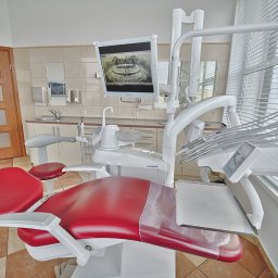 dento-sfera.pl WOJCIECH BILSKI - stomatolog, ortodonta - Czerwony fotel dentystyczny z widocznym zdjęciem panoramicznym zębów na monitorze i białymi roletami w tle.