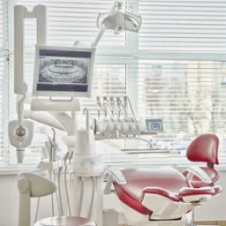 Nowoczesny gabinet dentystyczny z czerwonym fotelem, narzędziami i monitorem wyświetlającym zdjęcie rentgenowskie szczęki, oświetlony światłem dziennym wpadającym przez okno z żaluzjami.