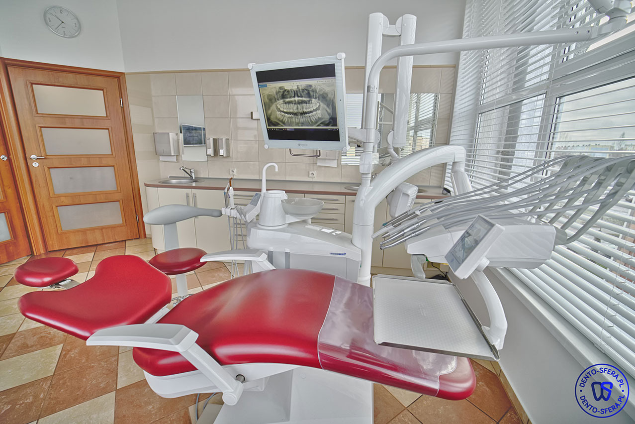 Czerwony fotel dentystyczny z widocznym zdjęciem panoramicznym zębów na monitorze i białymi roletami w tle.