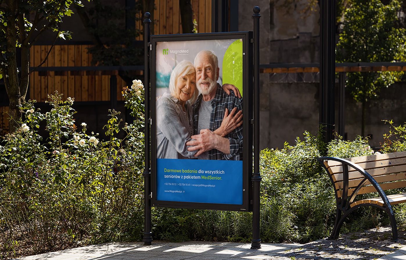 Billboard w parku z uśmiechniętą parą seniorów i reklamą MagnaMed: Darmowe badania dla seniorów. Ławka i zieleń w tle. Reklama usług medycznych.