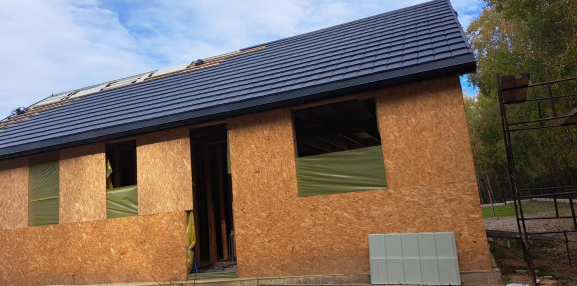 Dom w trakcie budowy z częściowo ukończonym dachem krytym ciemną dachówką, ściany zewnętrzne z płyt OSB, okna zabezpieczone folią, widoczne rusztowanie i drzewa w tle.