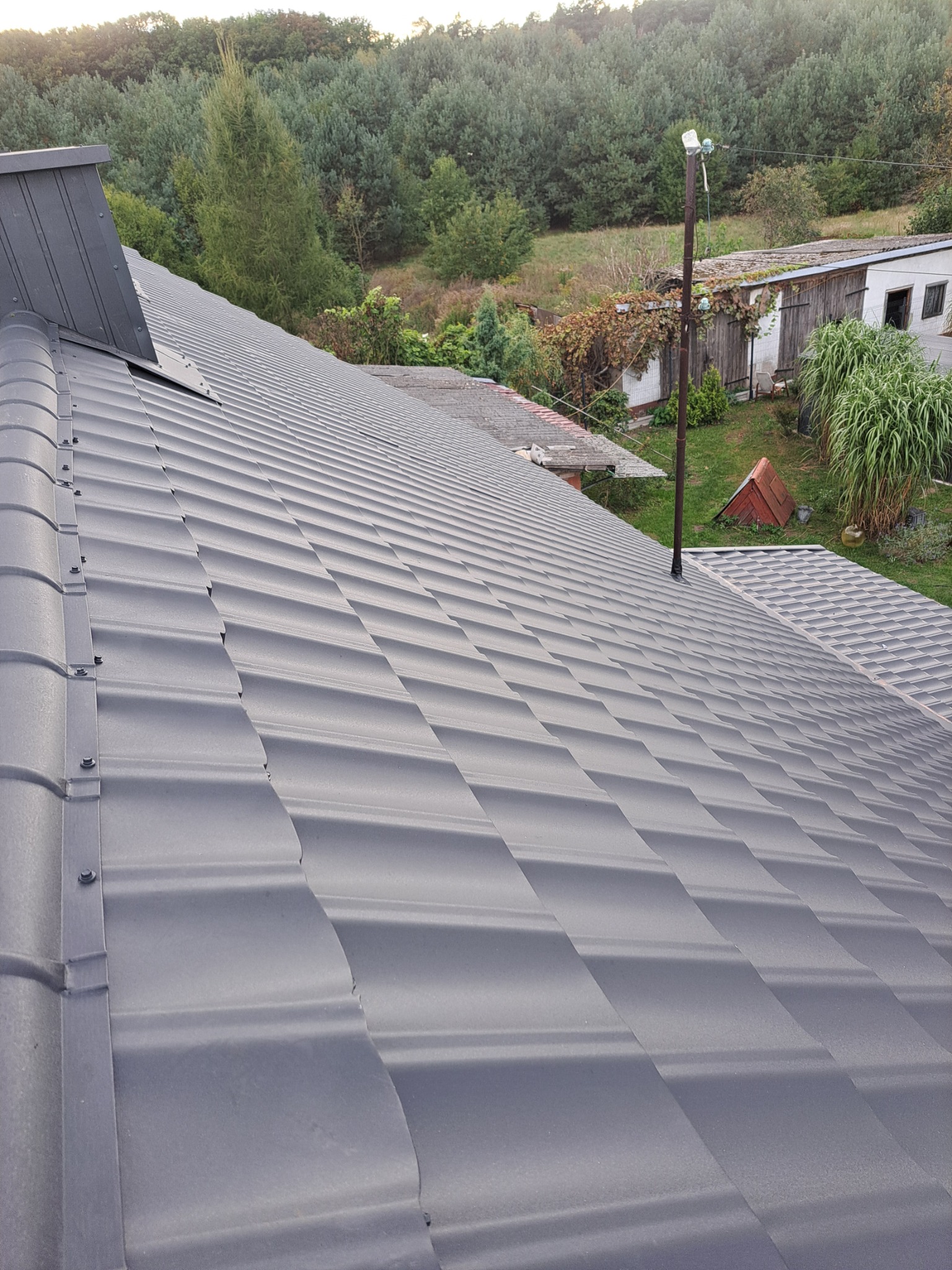 Szary dach z blachy trapezowej, widok z góry, w tle zieleń drzew i zabudowania gospodarcze.