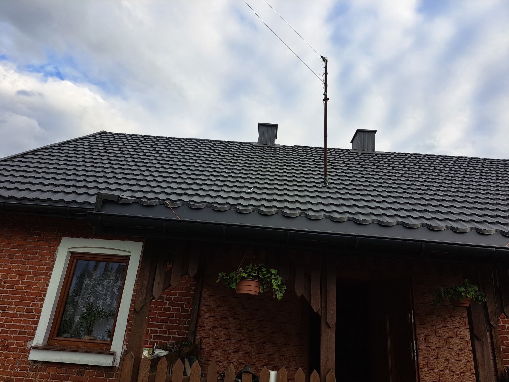 Dom z ciemnoszarą blachodachówką, widoczne kominy, rynny, instalacja odgromowa i wiszące doniczki z kwiatami.
