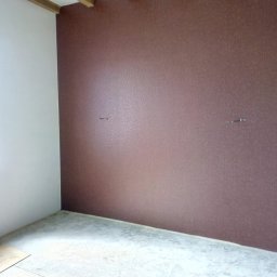 Svitlana REM - Pokój po remoncie: ściana w odcieniach brązu z delikatną fakturą, biała ściana boczna, surowa podłoga, drewniane belki sufitowe.