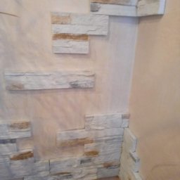 Svitlana REM - Fragment ściany wewnętrznej z dekoracyjnym wykończeniem z białych, nieregularnych płytek imitujących kamień, częściowo ułożonych na narożniku, na tle beżowej ściany z widoczną fakturą.