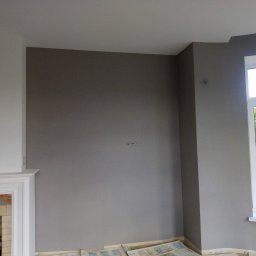 Svitlana REM - Świeżo pomalowane na szaro ściany pokoju z kominkiem, zabezpieczone podłogi gazetami. Widok na zewnątrz przez okno. Minimalistyczny design, spokojna kolorystyka.